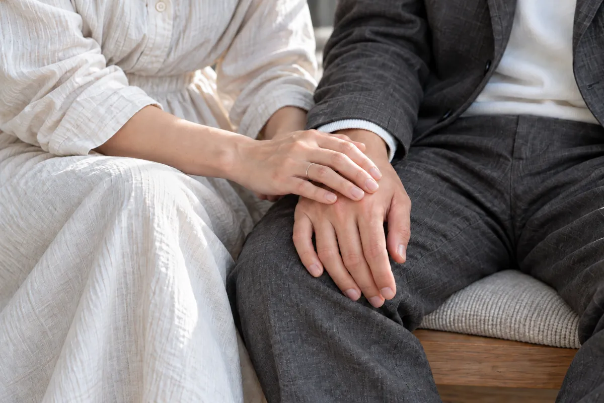 並んで座る夫婦。妻が夫の手にそっと手を重ねている手元のアップ